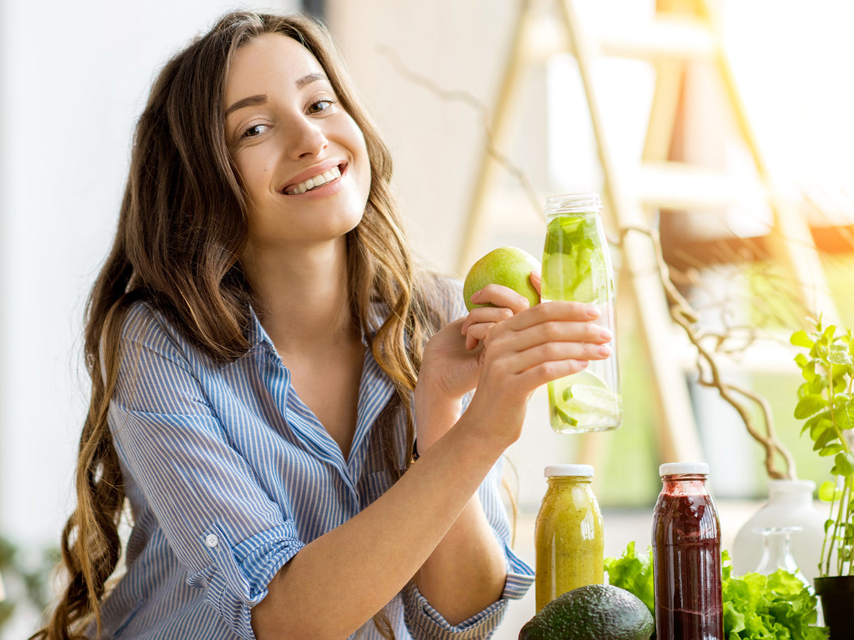 Young woman smiling while holding an apple and detox water, with fresh vegetables and juices on the table, highlighting a healthy lifestyle.