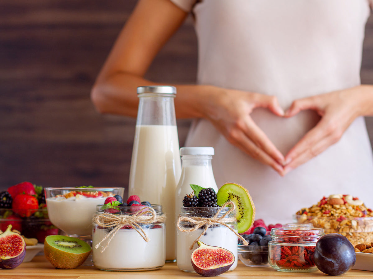 Close-up of nutritious foods like yogurt, berries, figs, kiwi, and grains, with a person forming a heart shape over their stomach, symbolizing gut and overall health.
