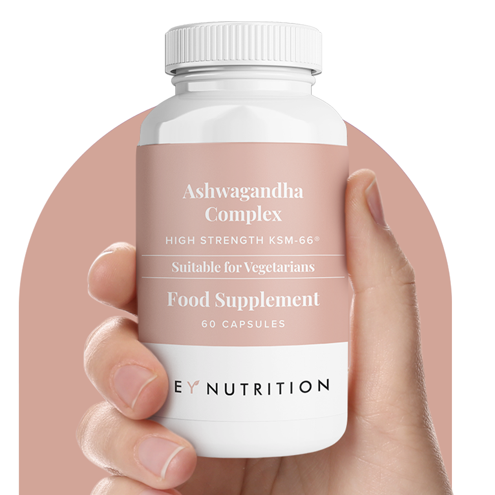 Hand holding Hey Nutrition Ashwagandha Complex supplement bottle with light pink label, pink background.