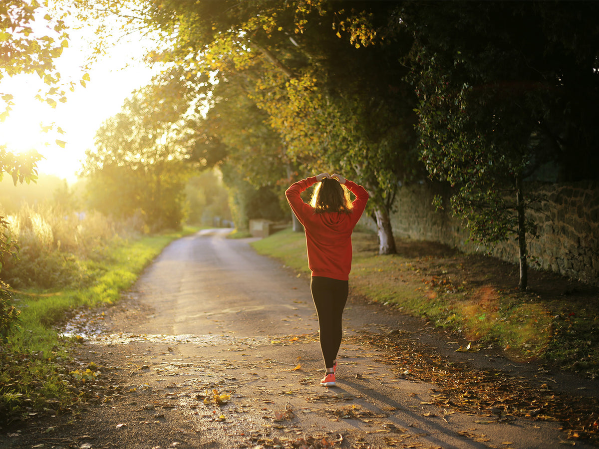 Woman walking outdoors on a sunlit path surrounded by trees, symbolizing overall wellness and vitality.