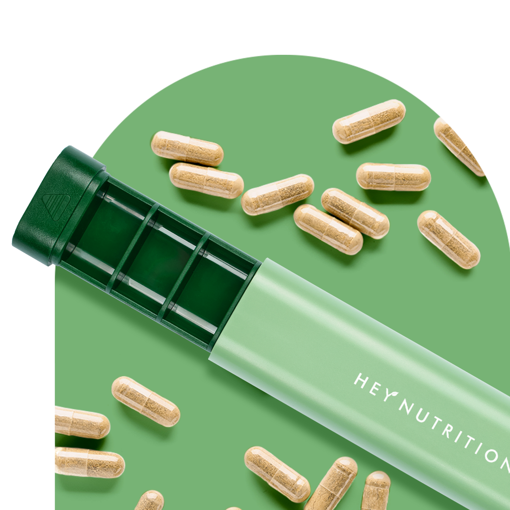 A green Hey Nutrition pill organizer box displayed with compartments visible, surrounded by several beige supplement capsules on a matching green background.