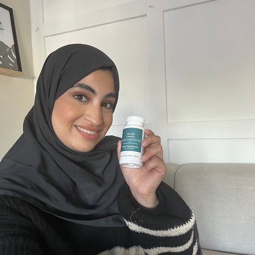 A woman wearing a black headscarf and striped sweater sitting on a sofa and holding Hey Nutrition Thyroid Complex, smiling warmly to showcase the supplement in a lifestyle setting.