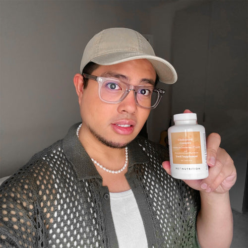 Portrait of a man wearing glasses and a cap, posing in a softly lit room while presenting a Hey Nutrition “Cholesterol Complex — Suitable for Vegetarians & Vegans — with Monacolin K & Added Plant Sterols — Food Supplement — 90 tablets” bottle.