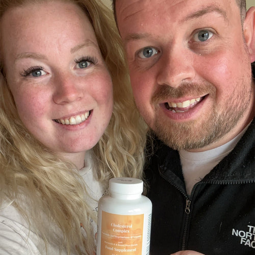 Close-up selfie of a smiling couple indoors holding a Hey Nutrition bottle labeled “Cholesterol Complex — Suitable for Vegetarians & Vegans — with Monacolin K & Added Plant Sterols — Food Supplement — 90 tablets,” front label facing camera.