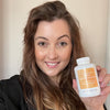 Bright indoor selfie of a woman in a black jacket smiling and holding a Hey Nutrition “Cholesterol Complex — Suitable for Vegetarians & Vegans — with Monacolin K & Added Plant Sterols — Food Supplement — 90 tablets” bottle beside her face.