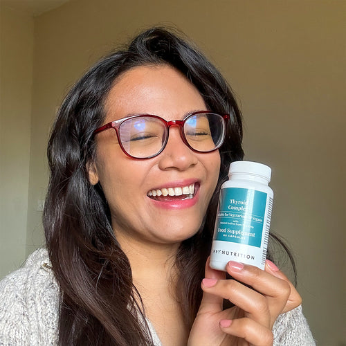 A woman with glasses and long dark hair laughing joyfully while holding a bottle of Hey Nutrition Thyroid Complex, conveying happiness and positivity associated with the product.