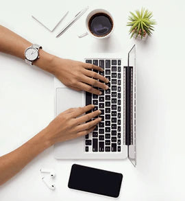 Top view of hands typing on a laptop keyboard, surrounded by a coffee cup, smartphone, notepad, pen, wireless earbuds, and a small potted plant on a white desk.