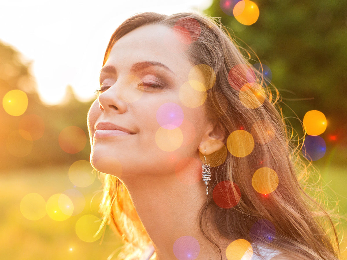Woman outdoors with soft sunlight and colorful bokeh effect, smiling peacefully with eyes closed, representing calmness and relaxation.