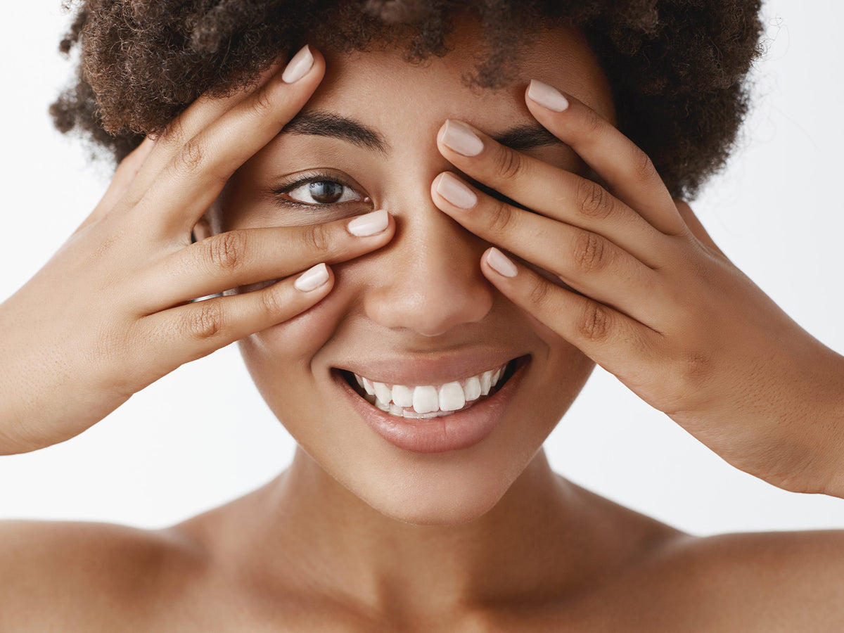 Smiling woman covering part of her face with hands – A close-up of a smiling woman with natural glowing skin, playfully covering her eyes with her hands, symbolizing wellness and vitality.
