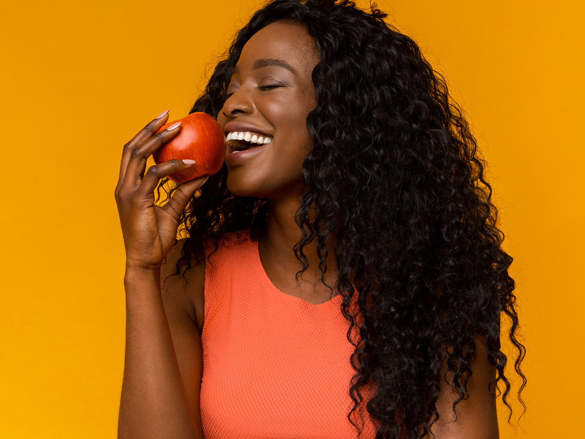 A smiling woman in an orange top holds a red apple close to her mouth against a bright yellow background.