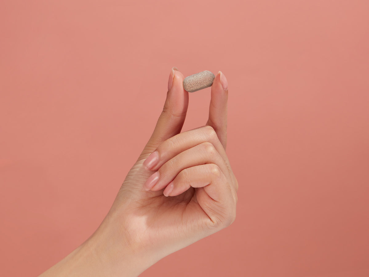 Hand gently pinching a small beige tablet against a soft peach background, illustrating the easy-to-swallow daily size of Hey Nutrition Cholesterol Complex designed to support healthy cholesterol and overall heart wellbeing.