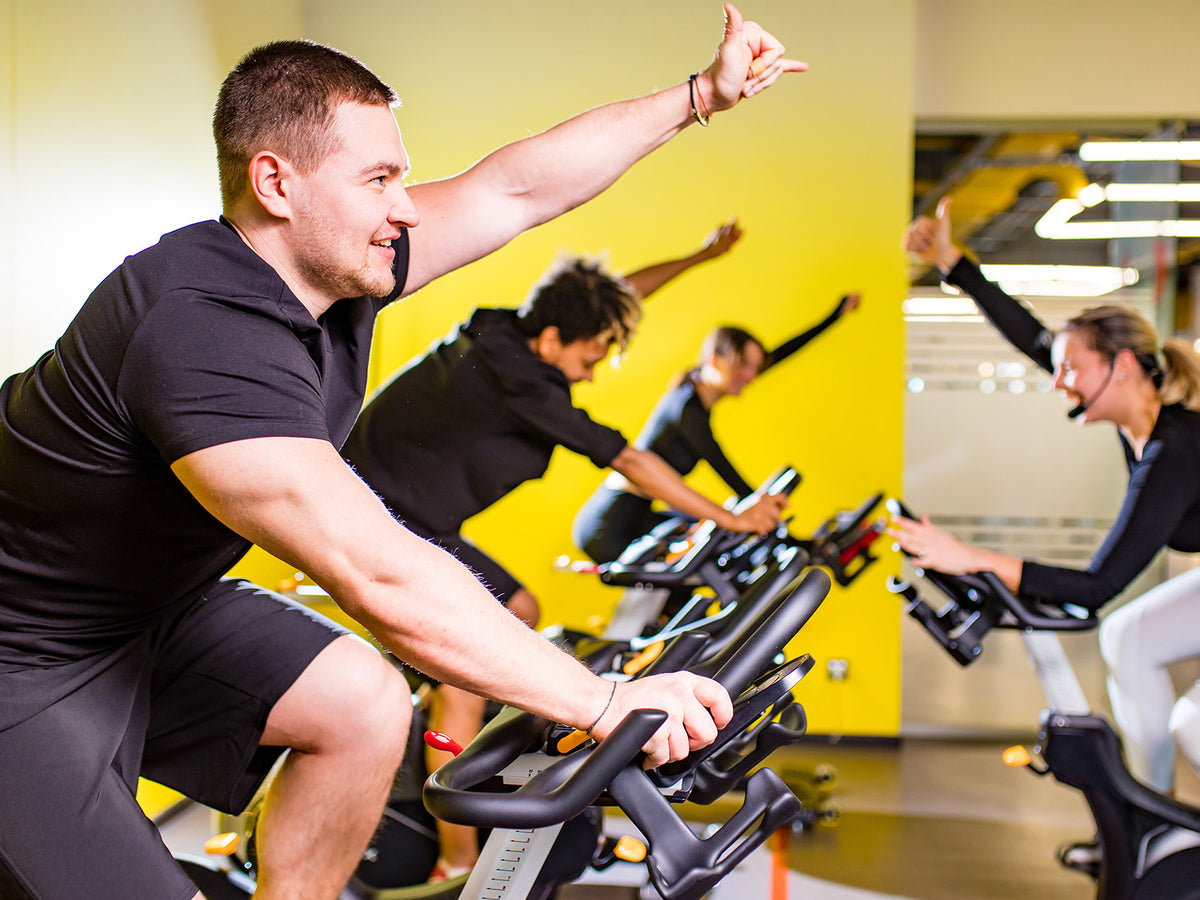 A group of people exercising together on stationary bikes in a brightly lit gym. The instructor and participants raise their arms in encouragement, representing energy and endurance.