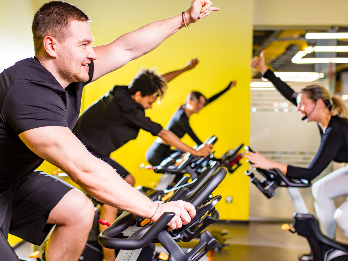 A group of people exercising together on stationary bikes in a brightly lit gym. The instructor and participants raise their arms in encouragement, representing energy and endurance.