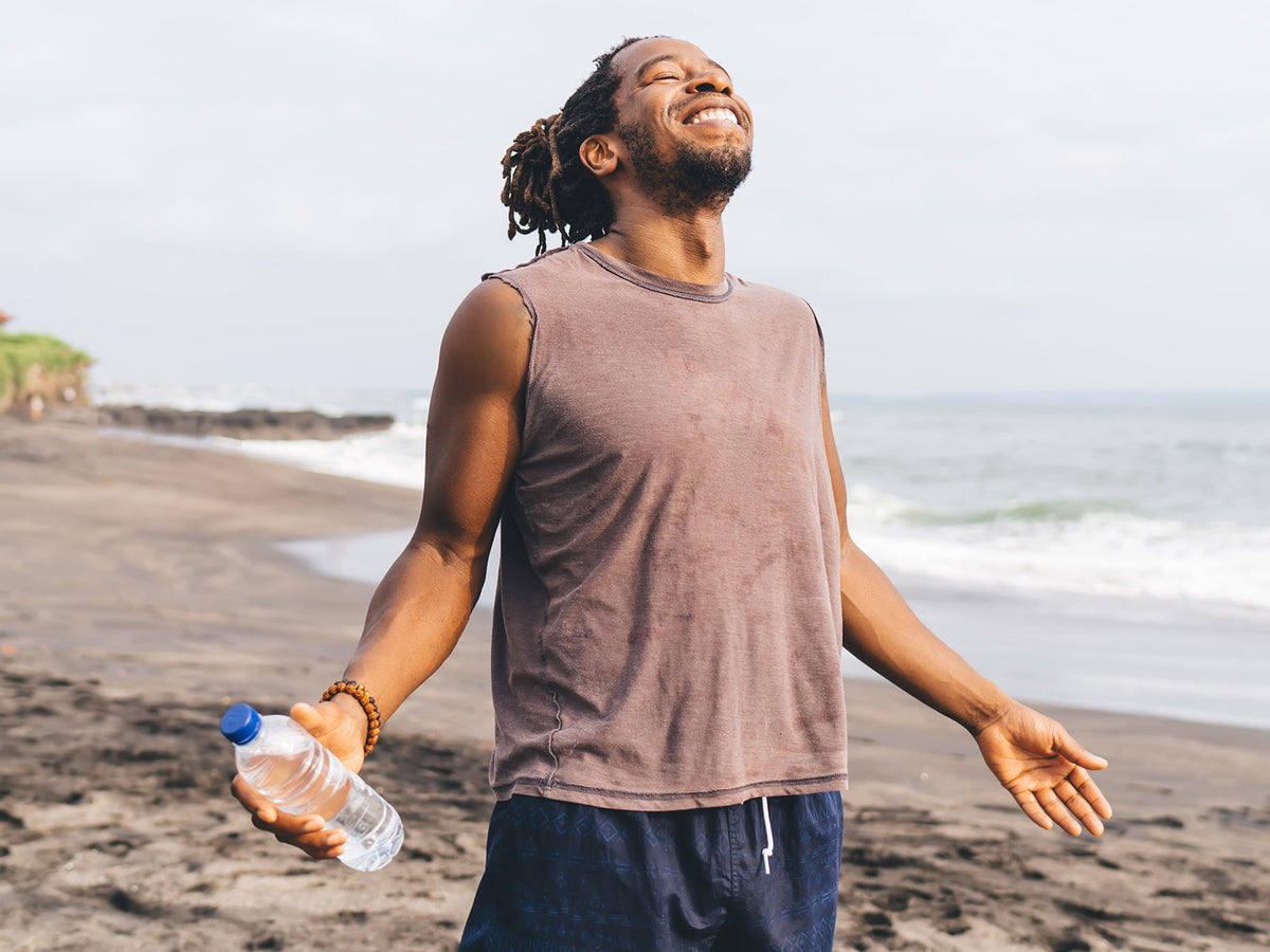 Happy man standing on the beach holding a water bottle, arms open and face lifted towards the sky, symbolizing energy, balance, and vitality.