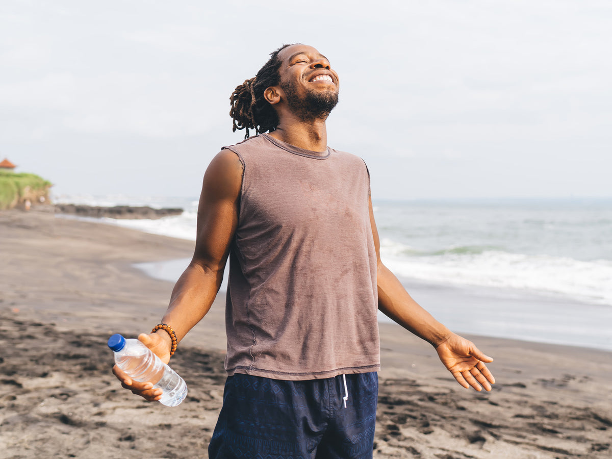 "Man standing on a beach holding a water bottle, arms outstretched and smiling, representing vitality, energy, and overall well-being supported by multivitamins and minerals."