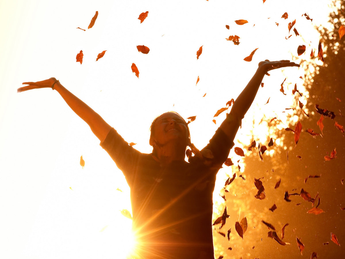 A woman standing in sunlight with arms raised, surrounded by falling autumn leaves, smiling with joy, symbolizing vitality and overall wellness.