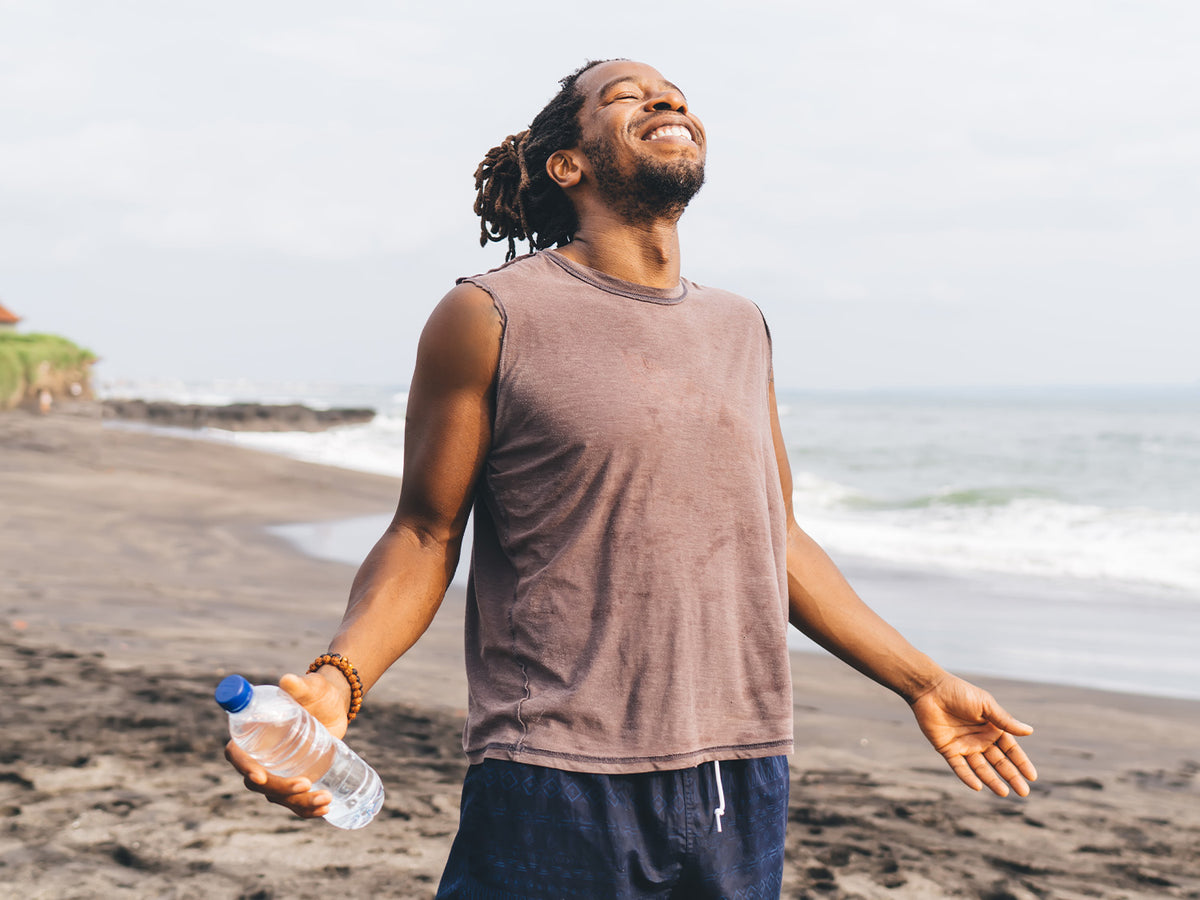 Happy man standing on the beach holding a water bottle, arms open and face lifted towards the sky, symbolizing energy, balance, and vitality.