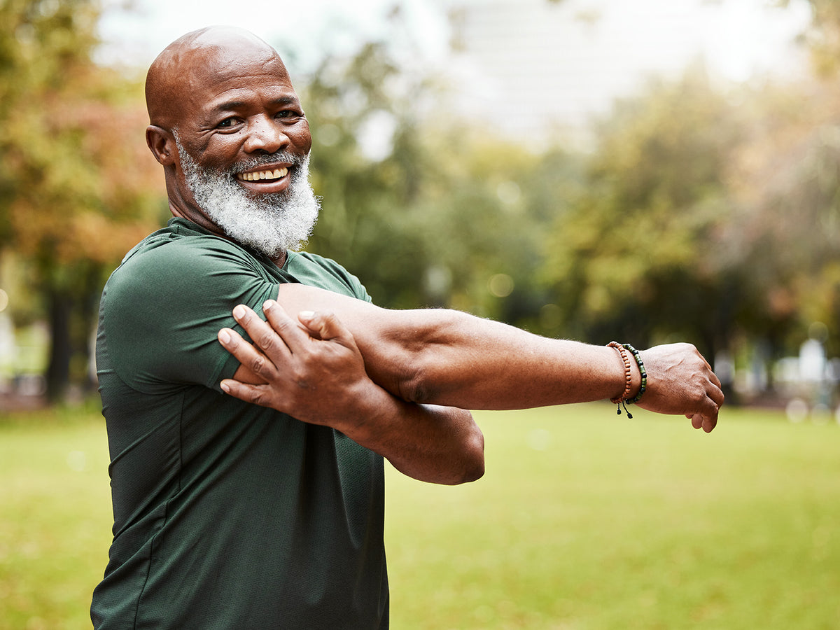 Smiling older man with a white beard stretching his arm outdoors in a park.