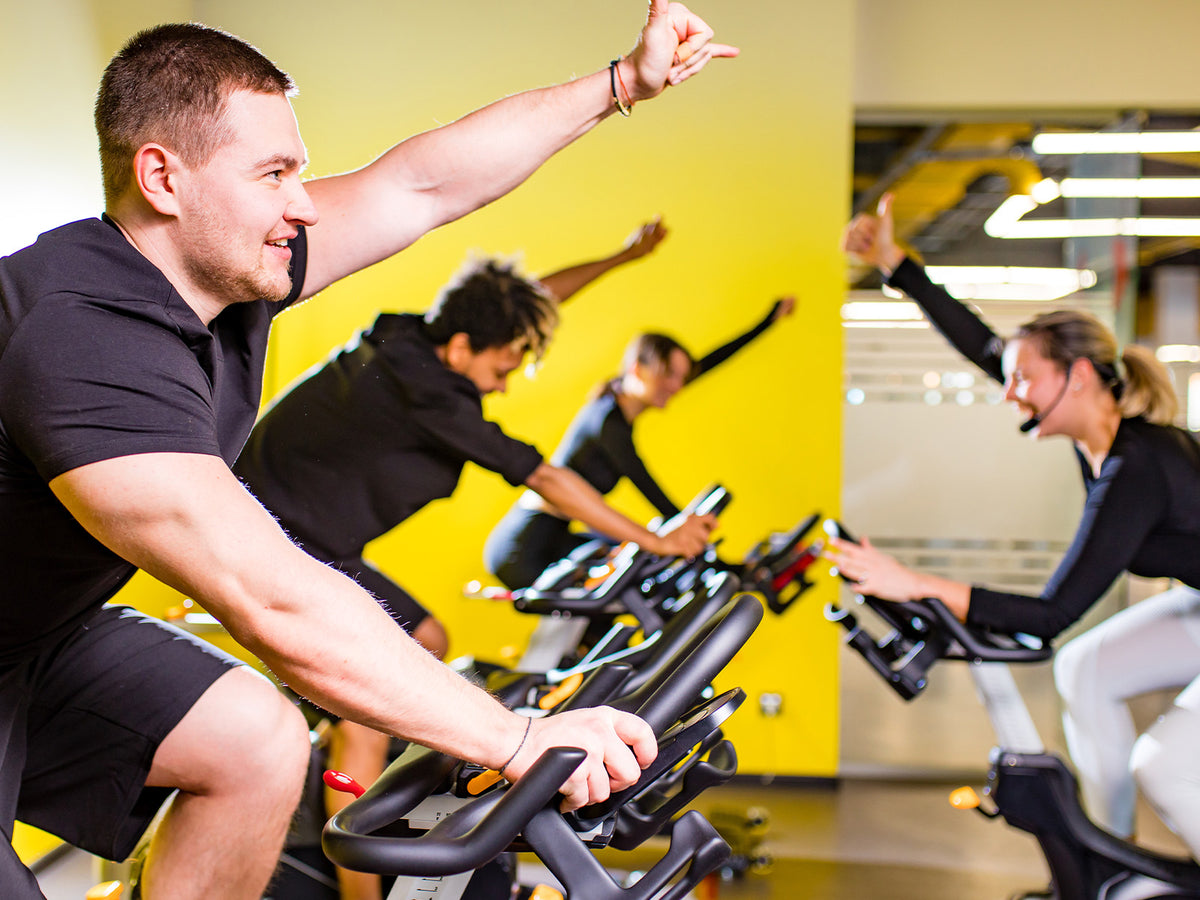 A group of people exercising together on stationary bikes in a brightly lit gym. The instructor and participants raise their arms in encouragement, representing energy and endurance.