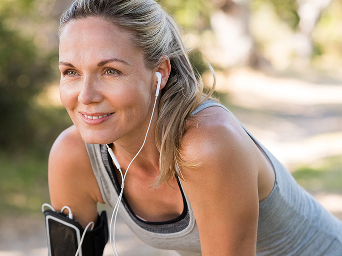 Woman in workout clothes smiling while exercising outdoors with earphones, promoting energy and vitality.