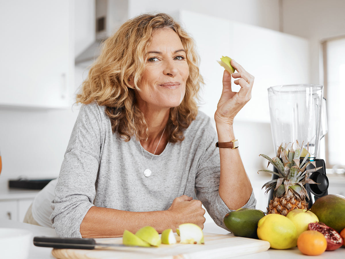 A smiling older woman in a kitchen eats a slice of apple with fresh fruits and a blender around her, representing healthy lifestyle choices for joint care.