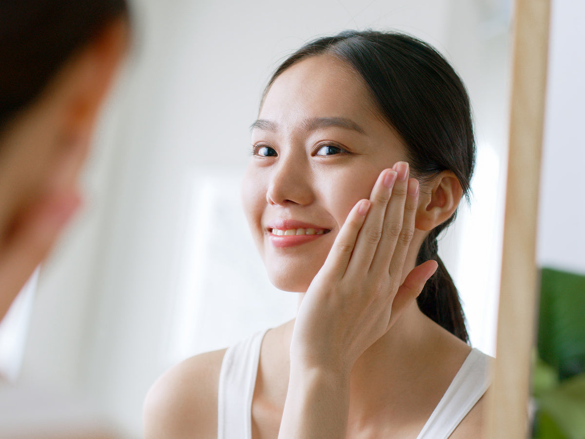 Woman smiling in mirror, gently touching her face with her hand, representing healthy glowing skin.