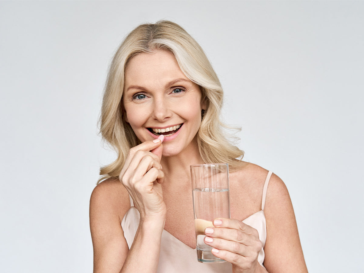 Mature woman smiling while holding a glass of water and a supplement capsule, promoting daily collagen support.