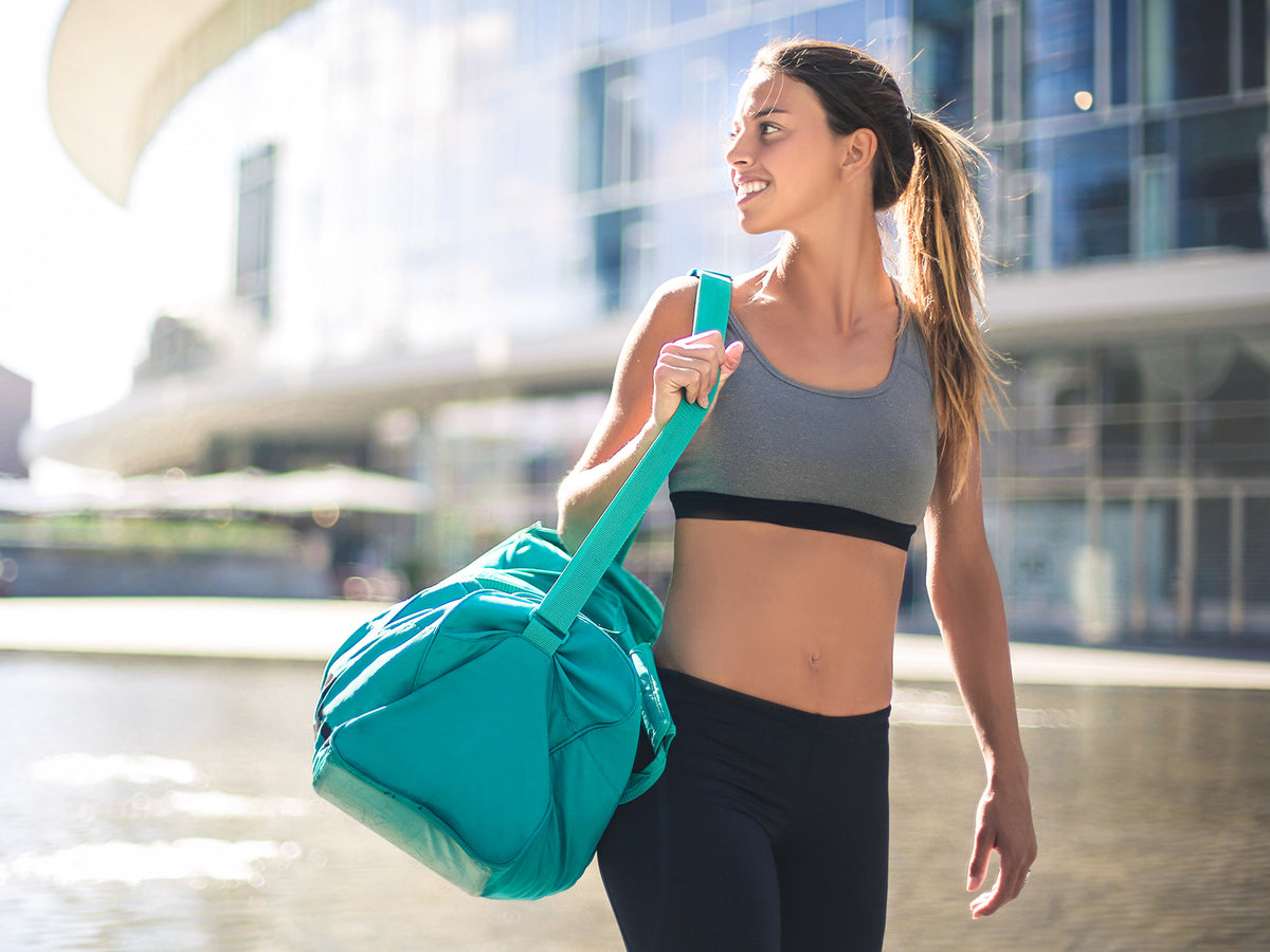 A fit young woman in a grey sports bra and black leggings carrying a turquoise gym bag, smiling while walking outside a modern glass building.