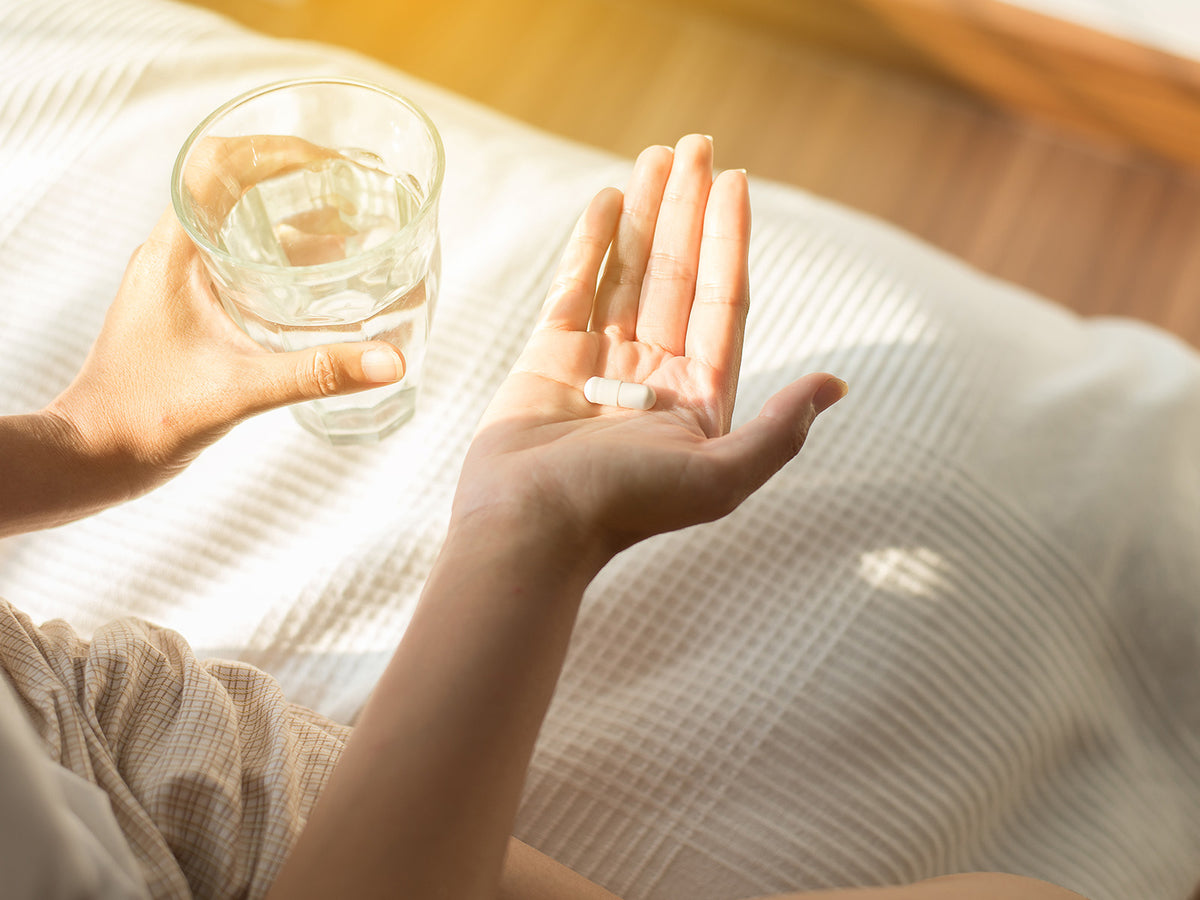 A person sitting in bed in soft morning light, holding a glass of water in one hand and a white capsule in the other hand, ready to take a supplement.