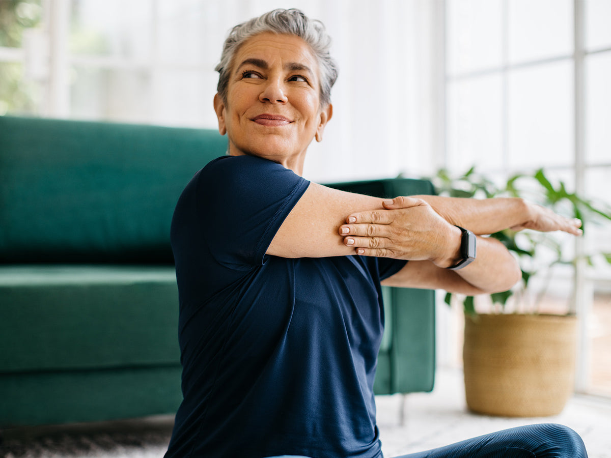 An older woman with short gray hair stretching her arm across her chest while sitting on the floor at home, smiling peacefully during a wellness or yoga session.