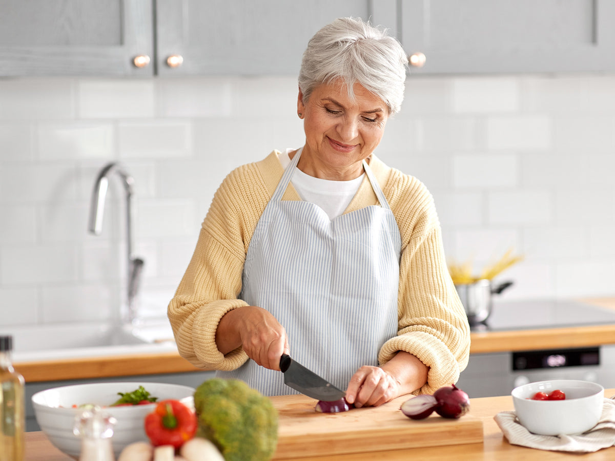 An elderly woman wearing a striped apron slicing a red onion in a modern kitchen, surrounded by fresh vegetables, smiling while preparing a healthy meal.