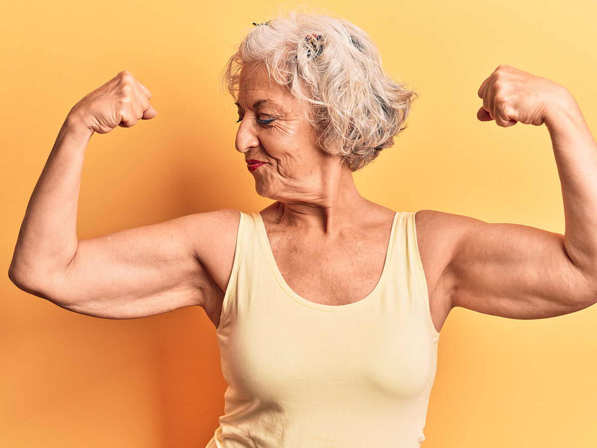 Strong older woman flexing both arms while smiling proudly, wearing a yellow tank top against a yellow background, representing strength, confidence, and healthy aging.