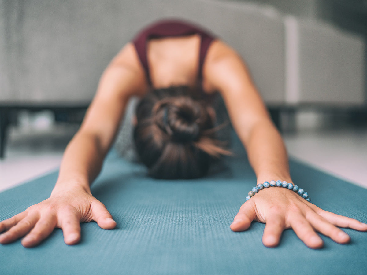 Woman practicing yoga in child’s pose on a teal mat, arms extended forward, focusing on calmness and flexibility in a peaceful indoor environment.
