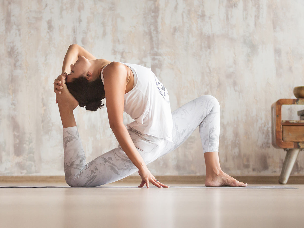 Woman in an advanced yoga pose with one leg bent behind her and held by both hands, demonstrating flexibility, balance, and concentration in a minimalistic room.