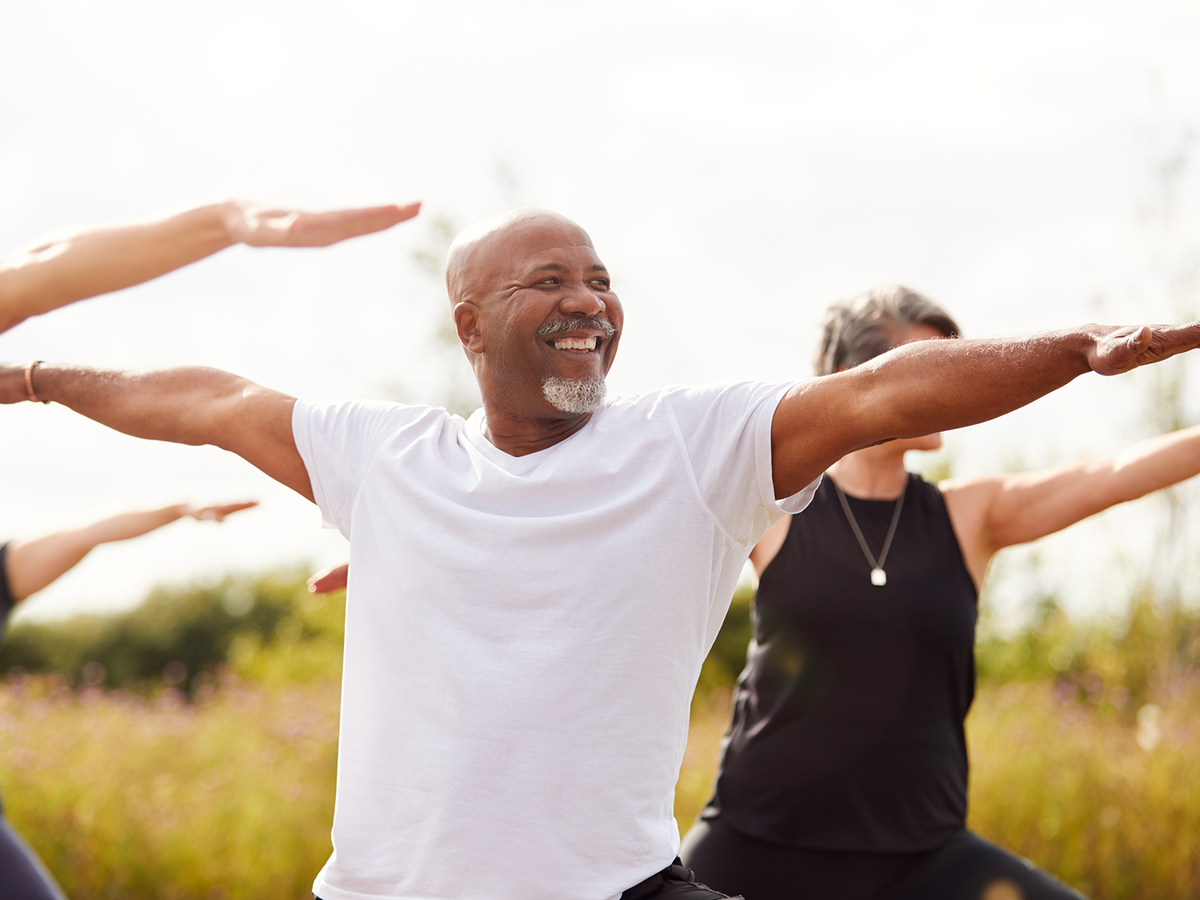 Smiling older man practicing yoga outside with a group, extending his arms in warrior pose in a grassy field under clear skies.