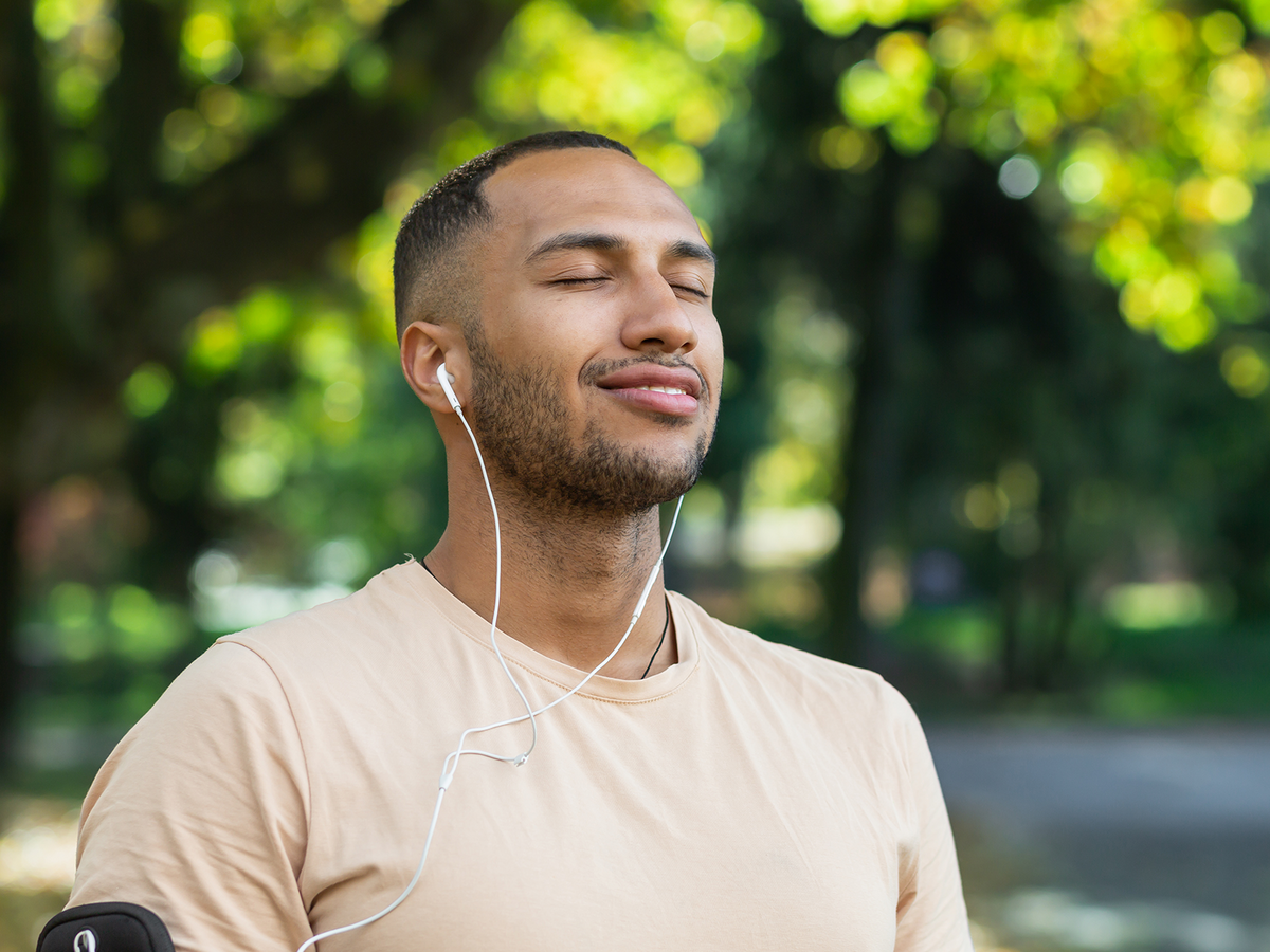 Young man with eyes closed and earbuds in, standing peacefully in a sunlit park while listening to music or a guided meditation.