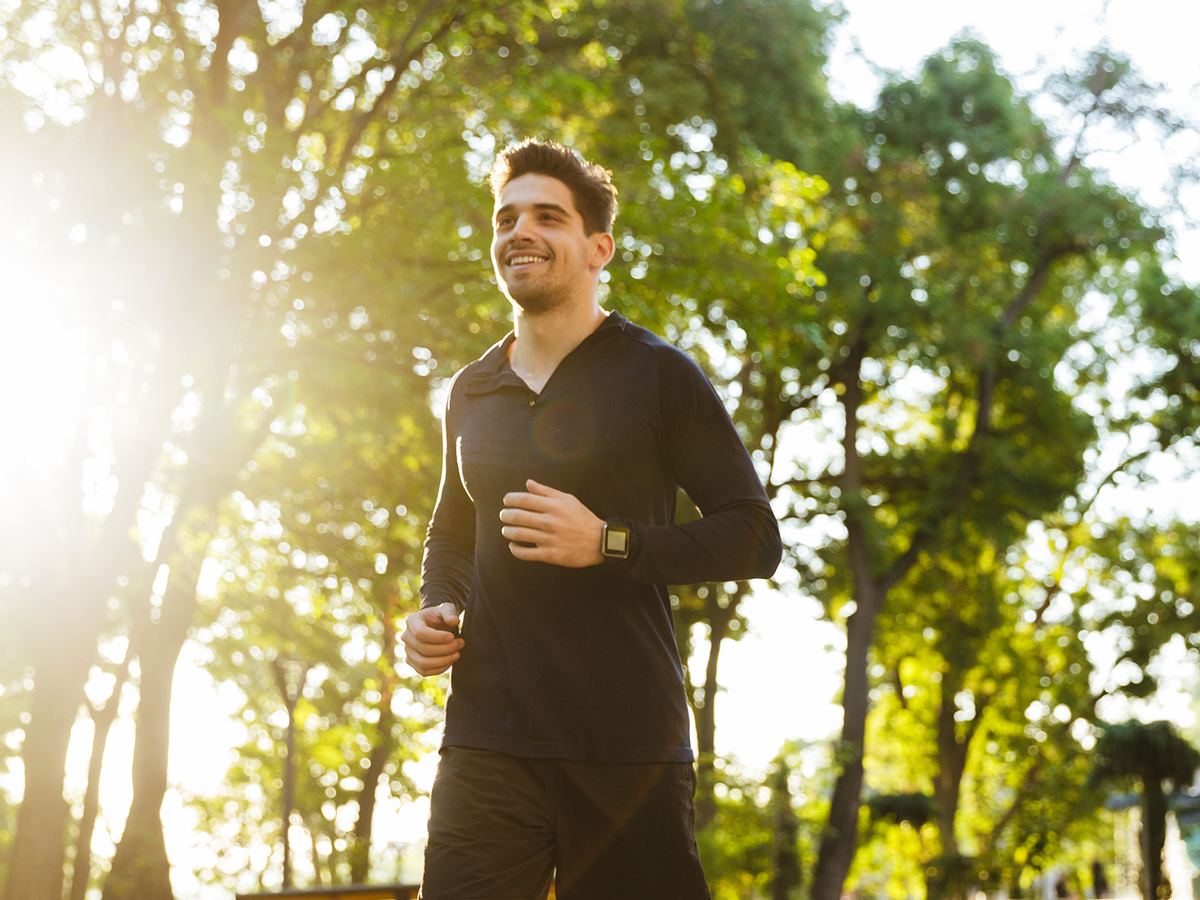 Young man jogging in a park surrounded by tall green trees, wearing athletic clothes and a smartwatch, with morning sunlight shining through.