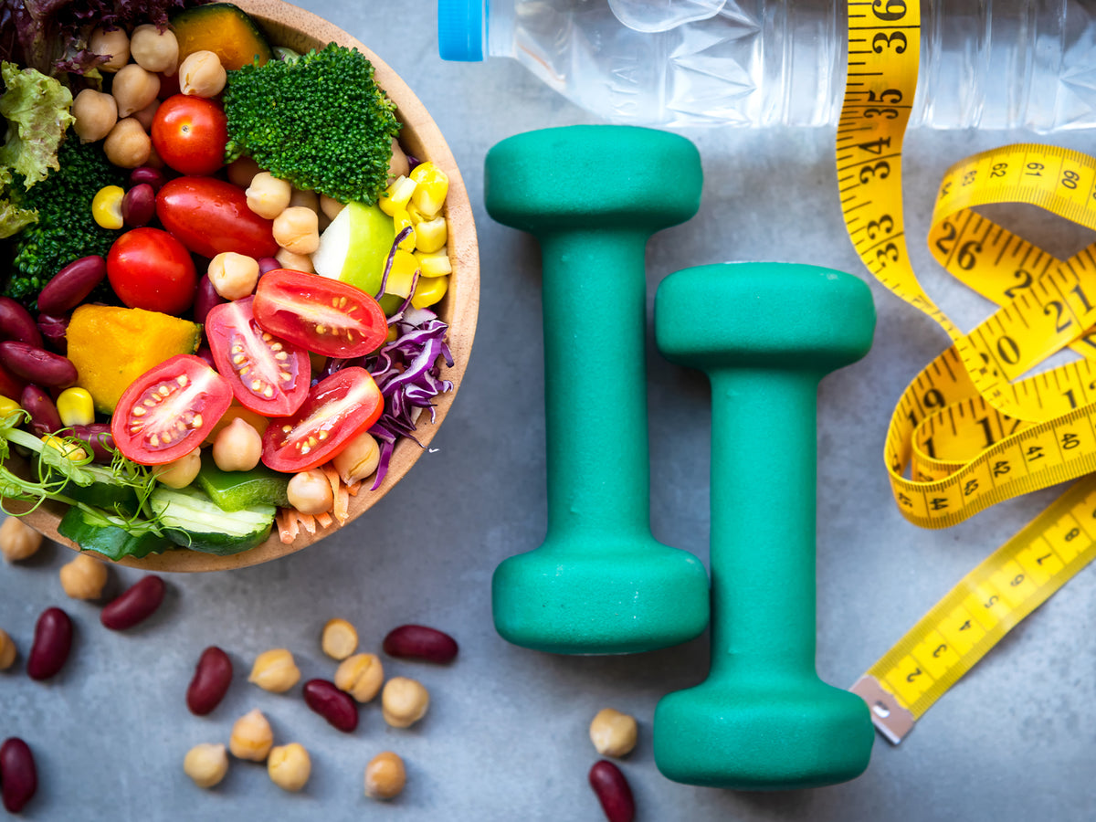 A health-focused flat lay image featuring a colorful salad with vegetables and legumes, two green dumbbells, a yellow measuring tape, and a water bottle—symbolizing fitness, nutrition, and weight management.
