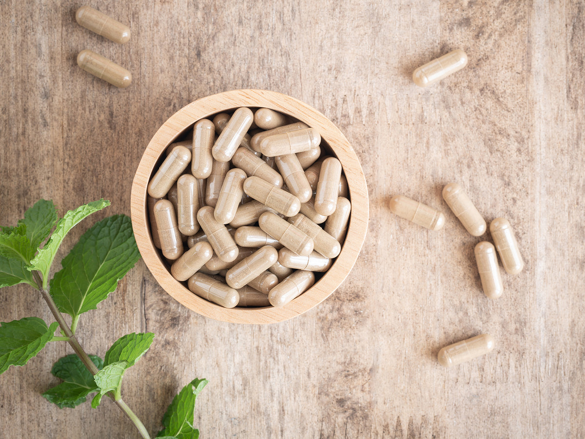 A top-down view of a wooden bowl filled with beige herbal supplement capsules, surrounded by scattered capsules and fresh mint leaves on a wooden surface—evoking natural, plant-based supplementation.